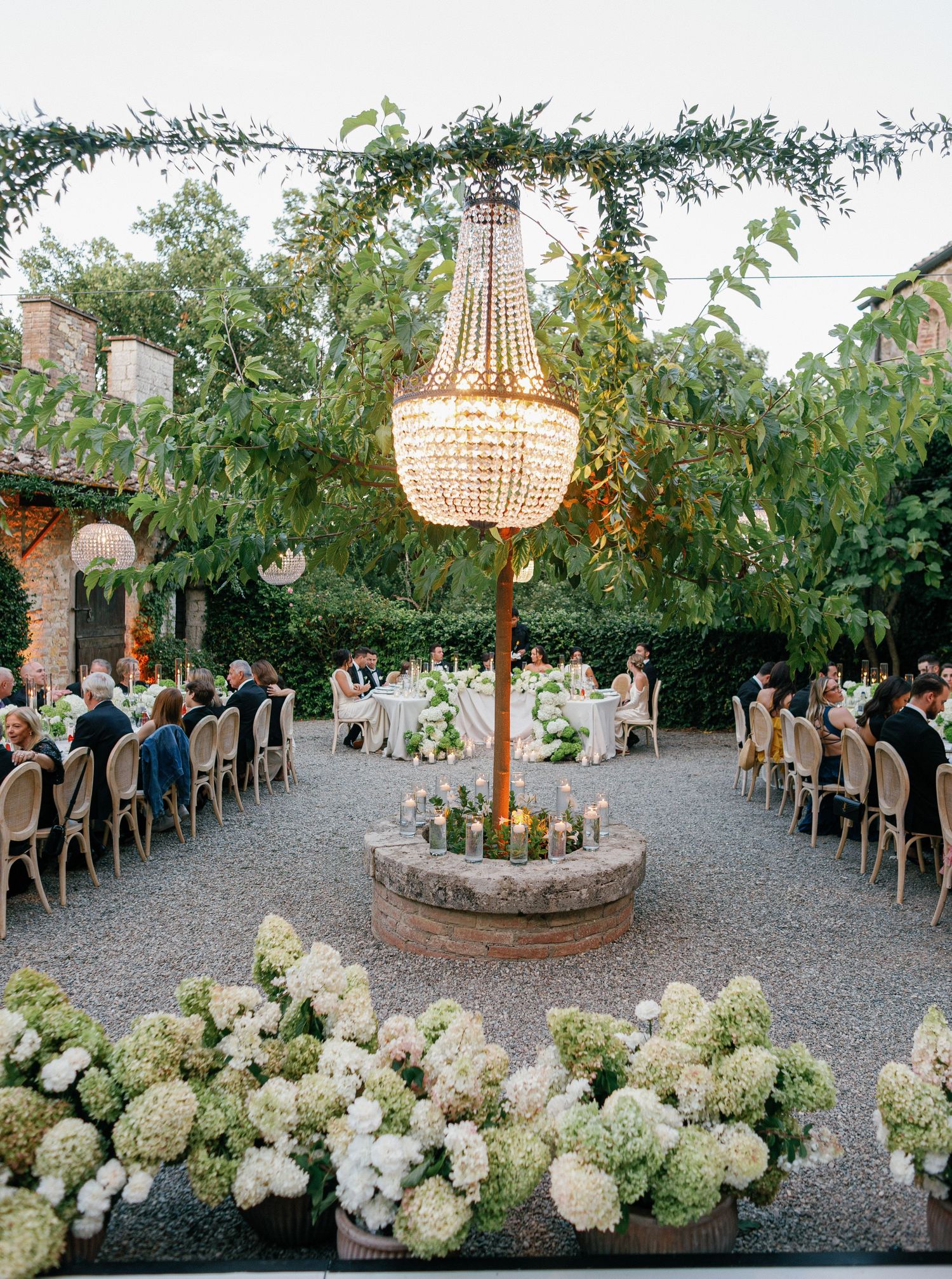 Panoramic view of outdoor wedding reception with crystal chandelier, gravel path, and white floral arrangements.