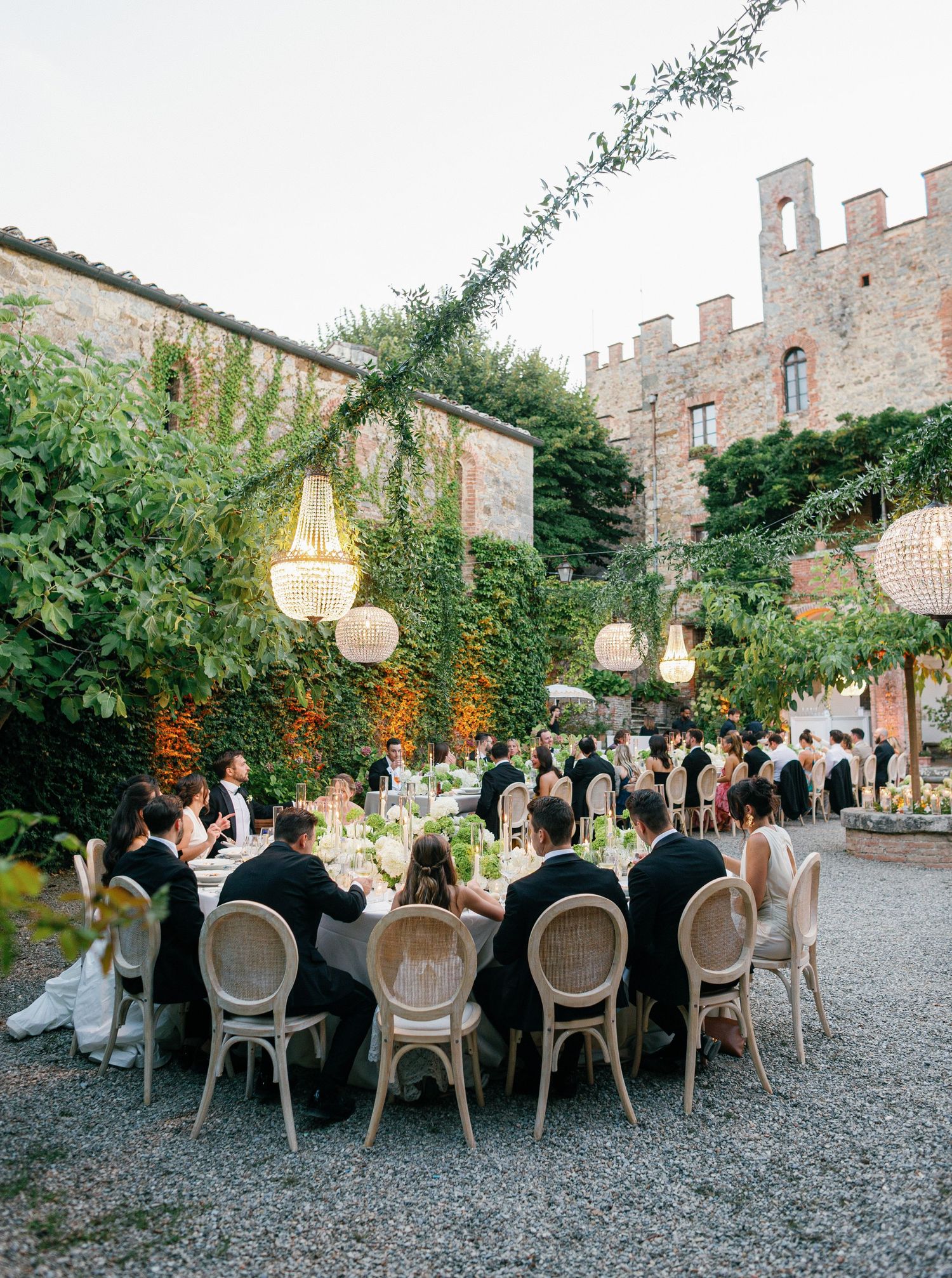 Elegant outdoor wedding reception dinner setup with long tables, classic chairs and hanging lanterns at a stone castle venue.