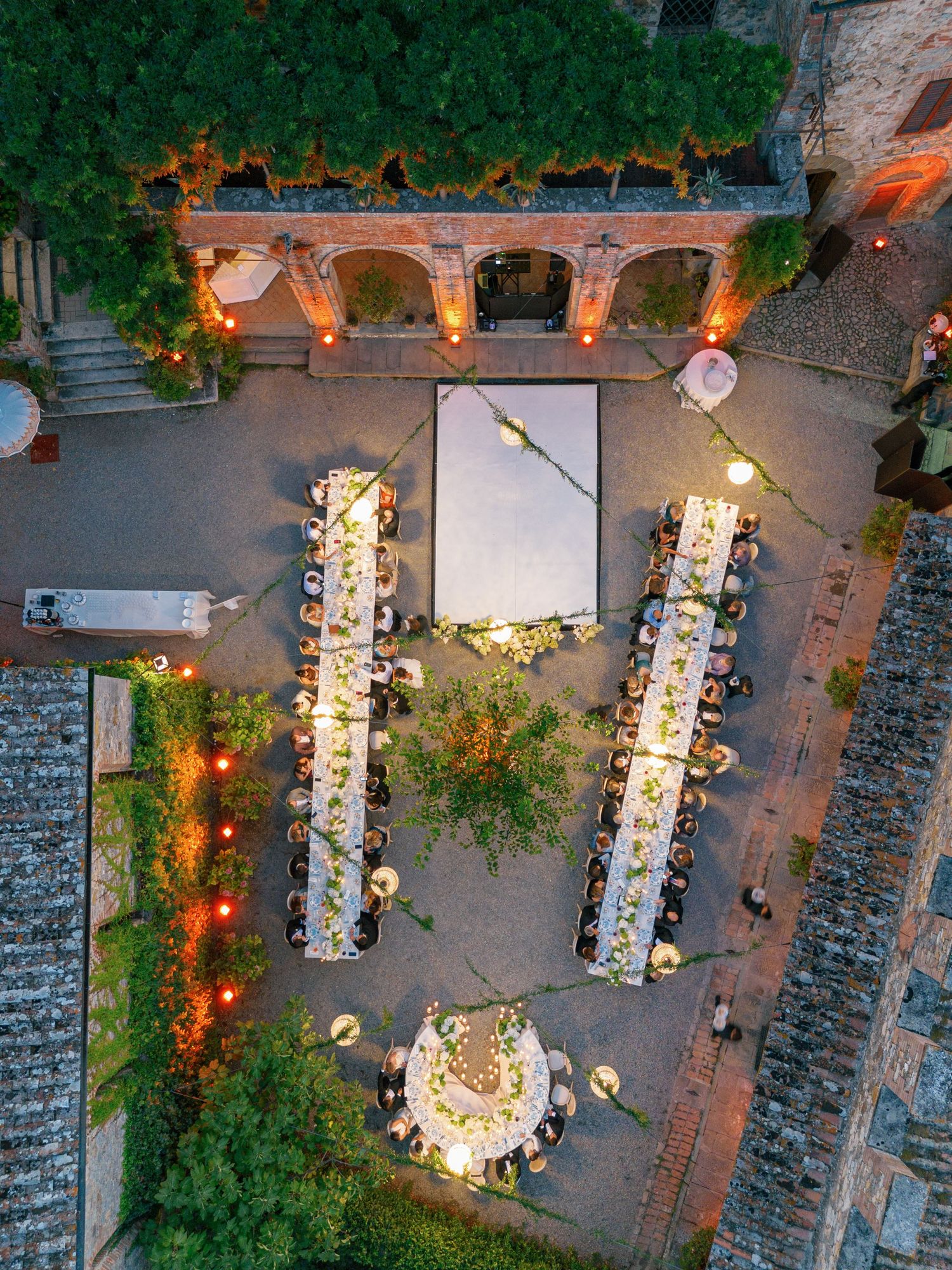 Aerial view of an elegant outdoor wedding reception with long tables and string lights under stone archways at dusk.