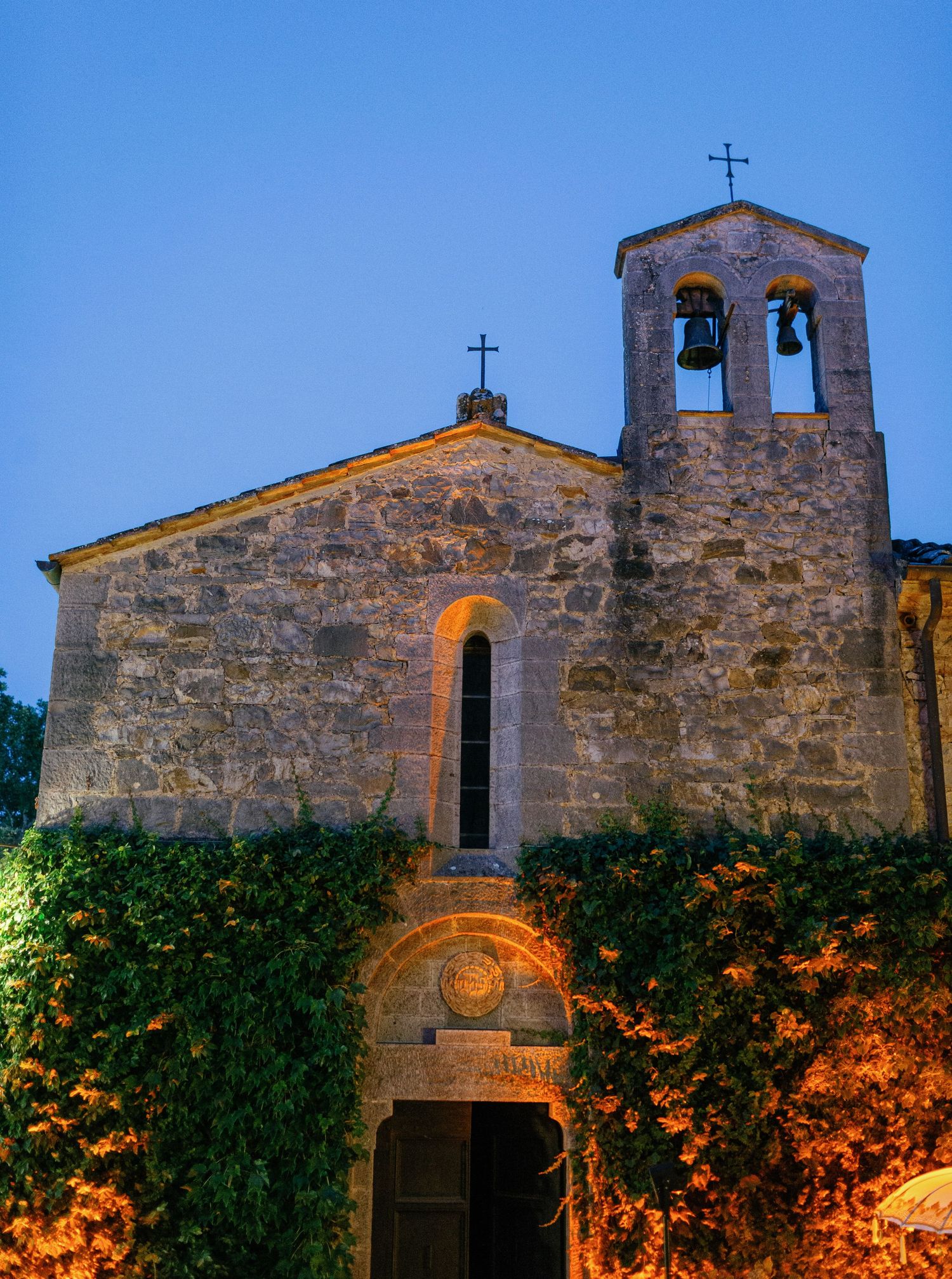 Ancient stone church with bell tower illuminated at dusk, with climbing vines and warm lighting around entrance.