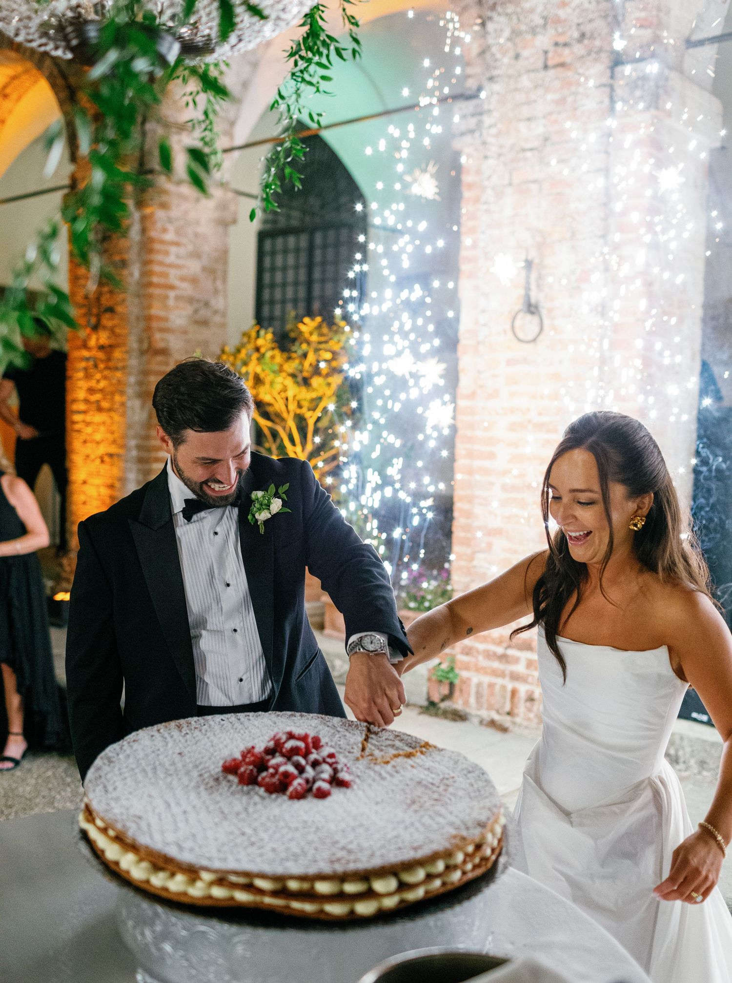 Couple signs their wedding guestbook while string lights twinkle against exposed brick walls at a rustic venue.