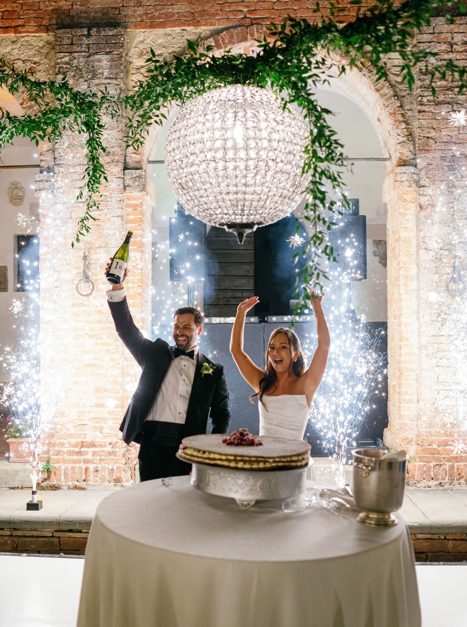 A joyful couple celebrates under elegant crystal chandeliers and greenery at a rustic brick venue reception.