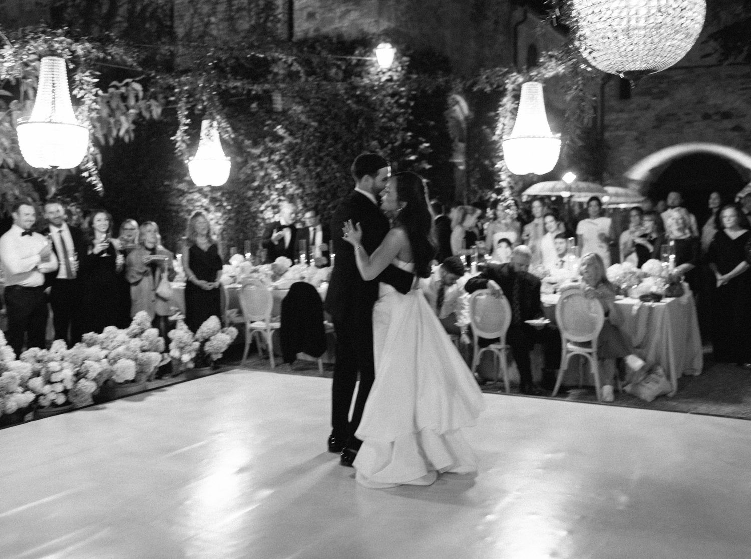 A couple shares their first dance at a wedding reception under glowing lights while guests watch from their tables.
