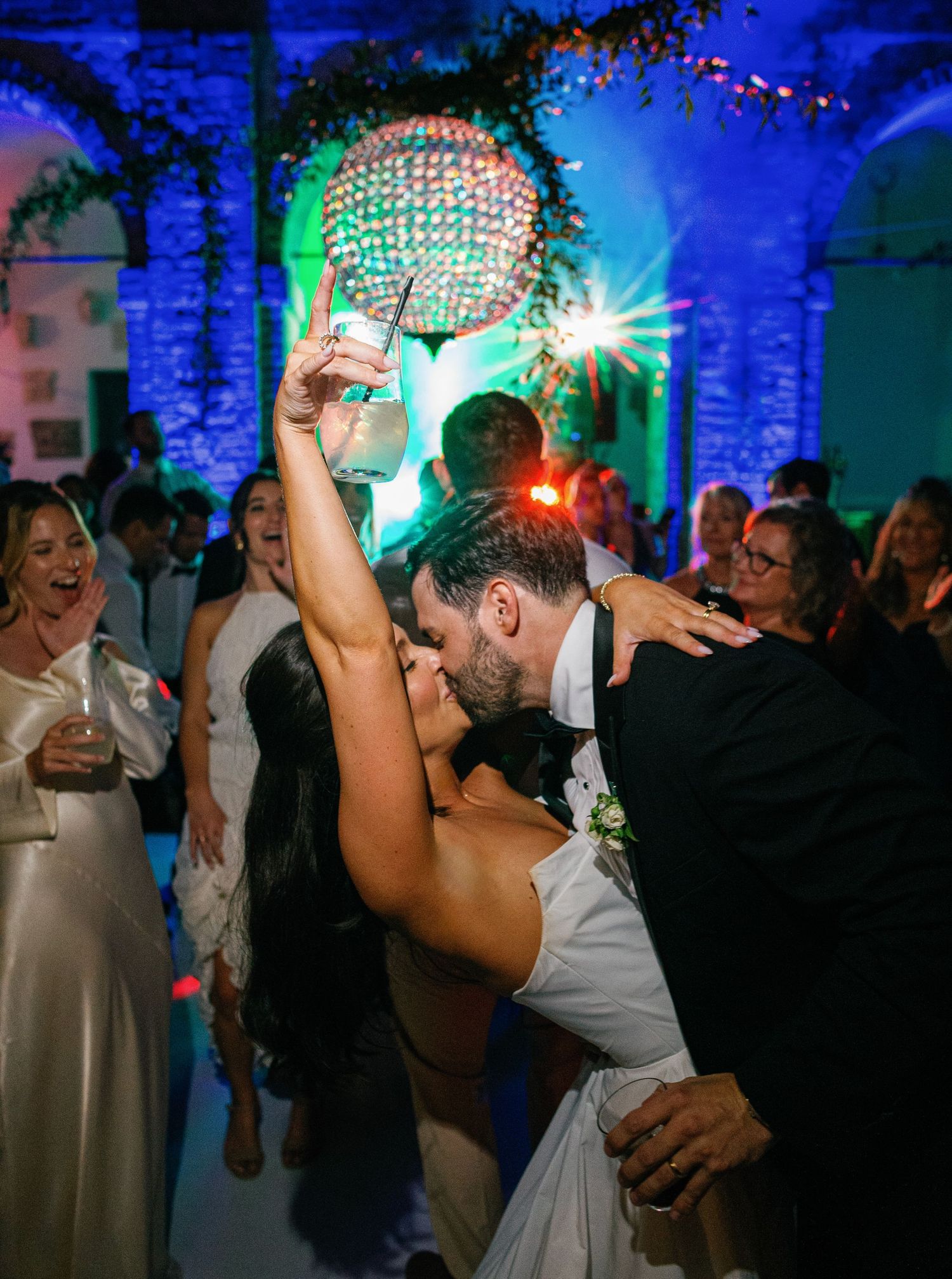 Wedding couple and guests dance joyfully under blue lighting and festive decorations at an evening reception celebration.