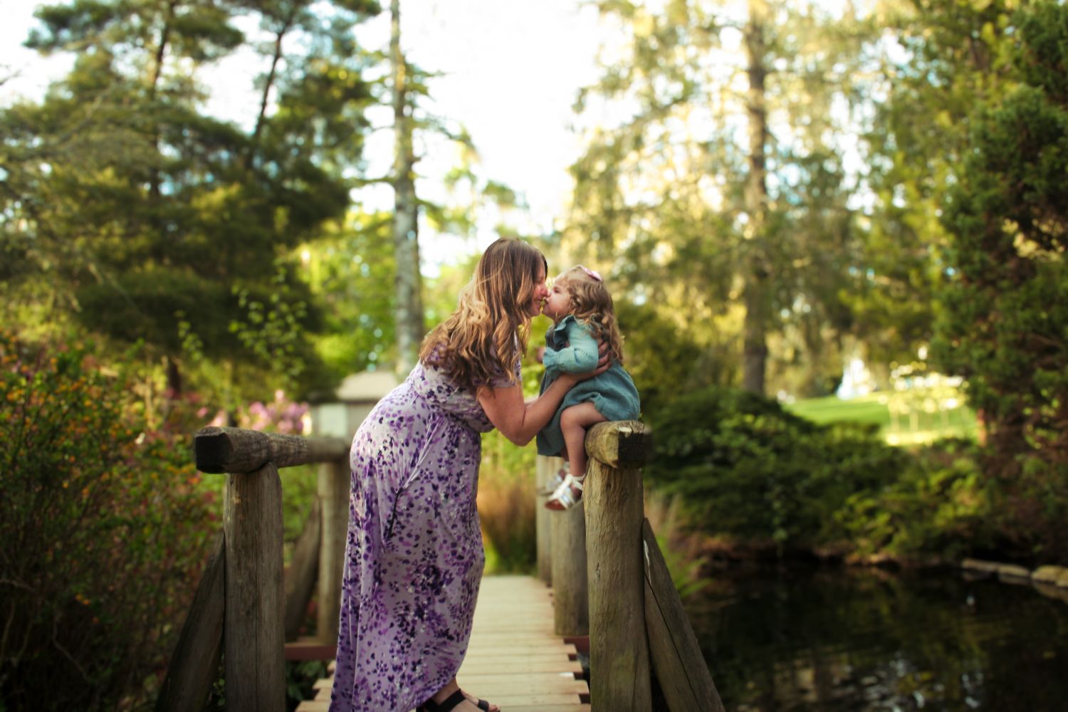 A couple walks hand in hand along a sunlit wooden bridge surrounded by lush green trees and foliage.
