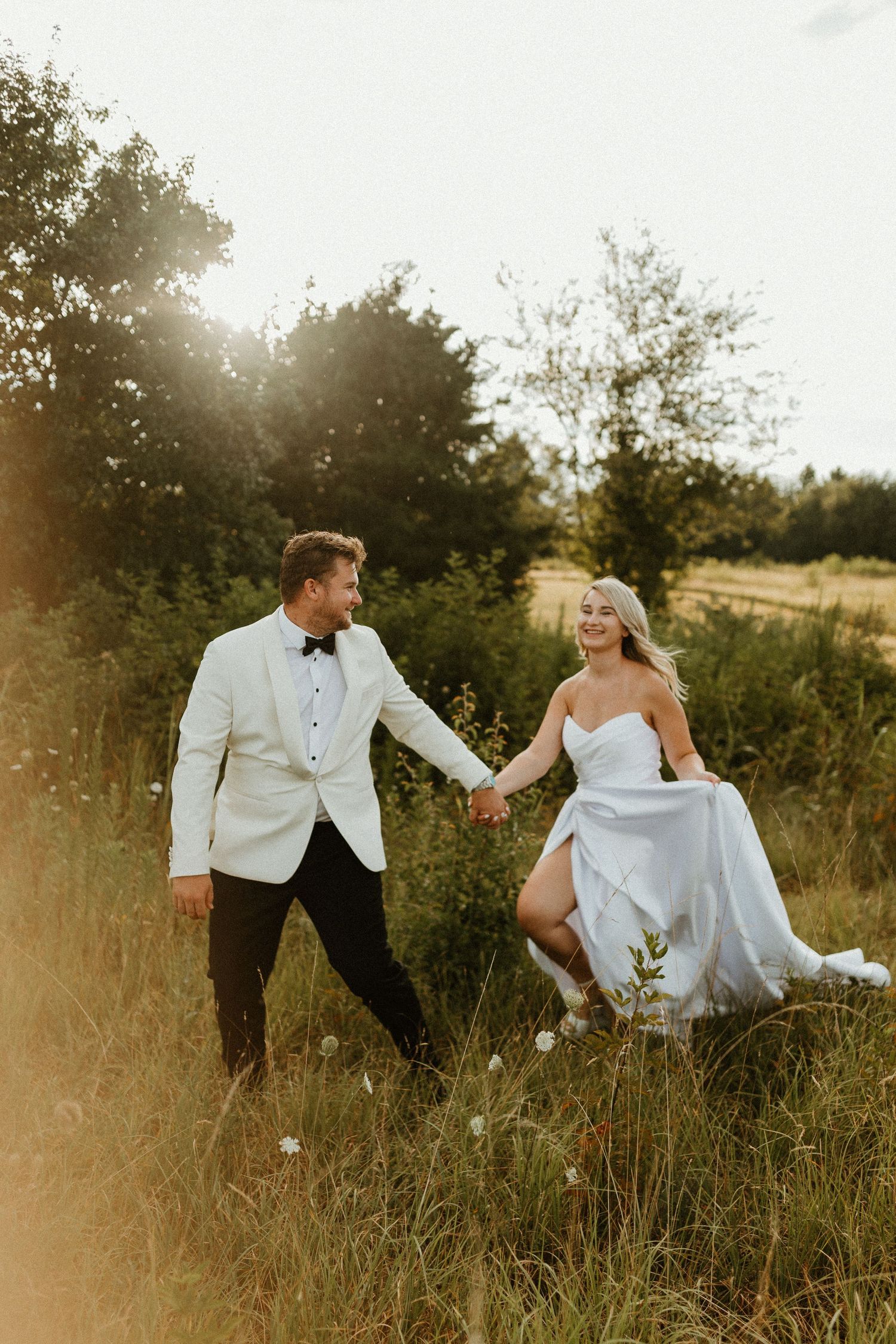 A couple in wedding attire playfully poses in a sunlit meadow during golden hour.