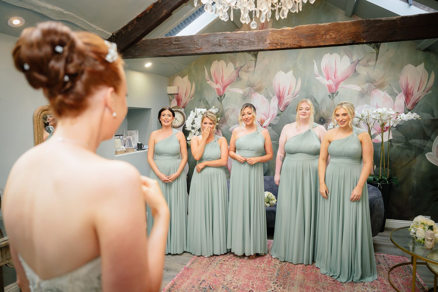 Bridesmaids wearing matching sage green dresses stand together in an elegant venue with a chandelier. Woodhill Hall.