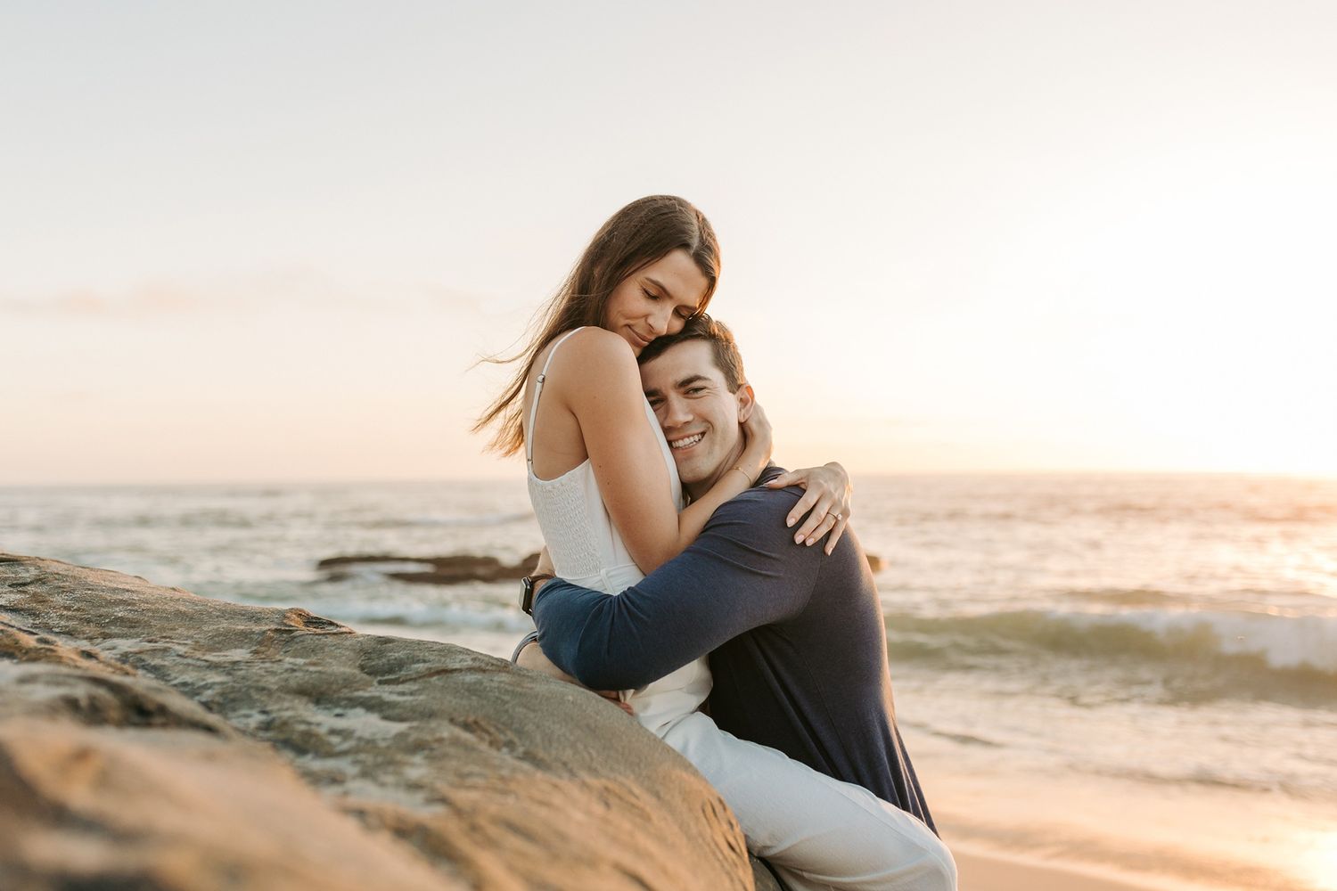 A couple embraces on a sandy beach during golden hour as waves crash behind them in a romantic seaside setting at Windansea Beach.