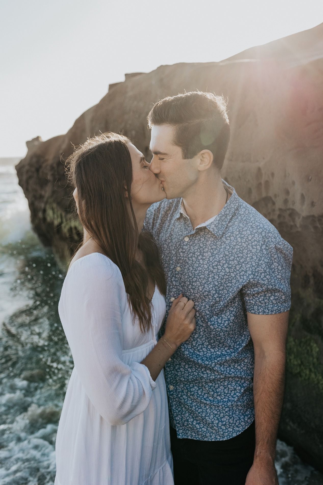 A couple in white dress and blue floral shirt share romantic moments against a sunlit ocean backdrop at Windansea Beach.