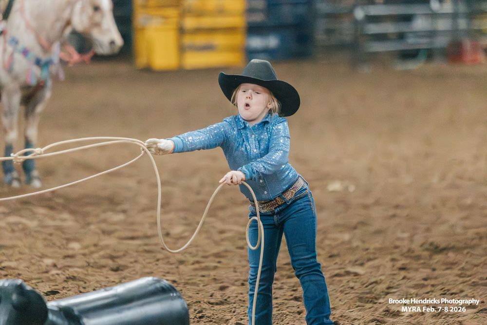 Young cowboy in blue western attire practices roping skills in a dusty rodeo arena.