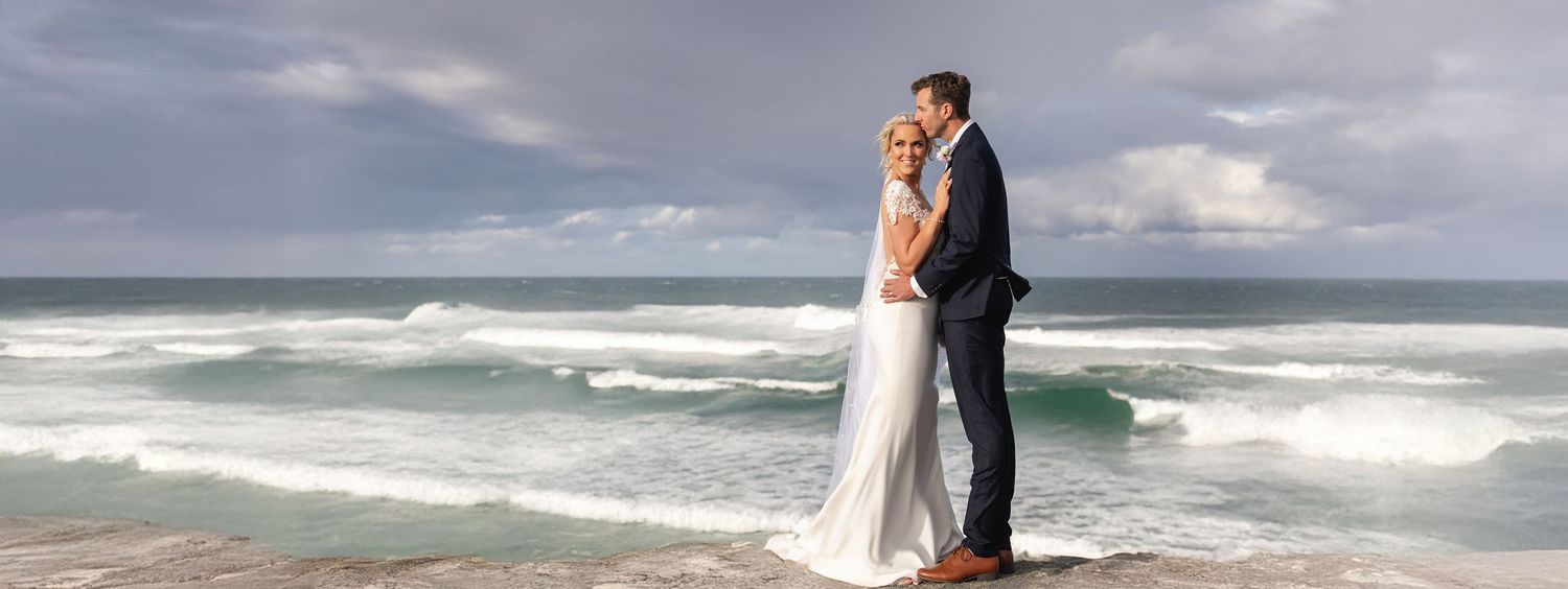 Newlywed couple share a tender kiss on seaside rocks with turquoise waves crashing behind them.