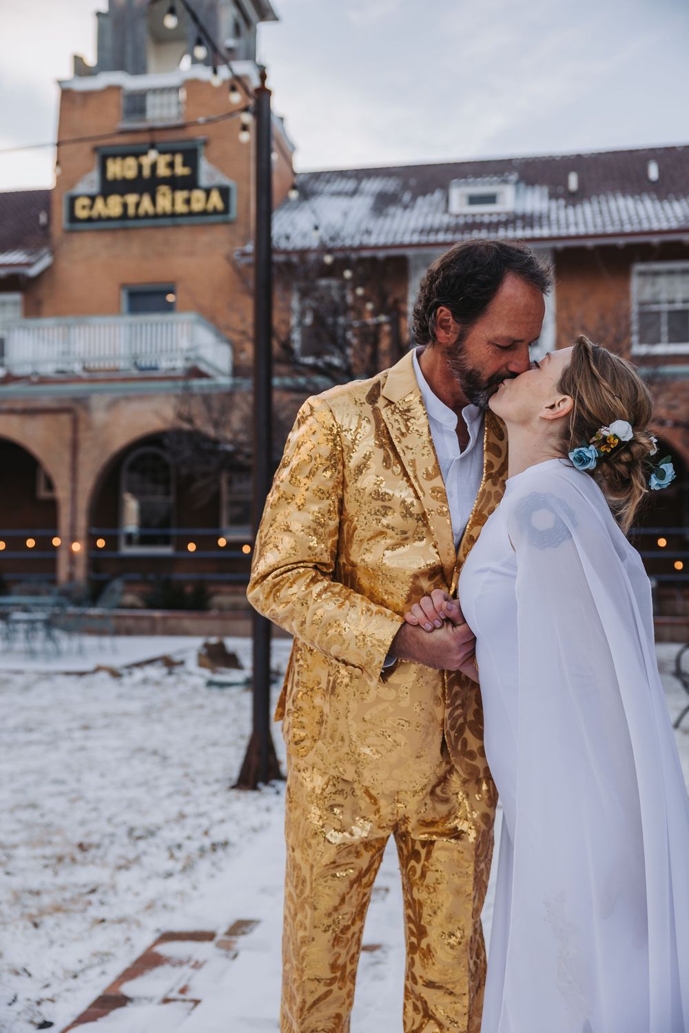 A couple in a gold suit and white dress share an intimate moment outside Hotel Castaneda in winter.