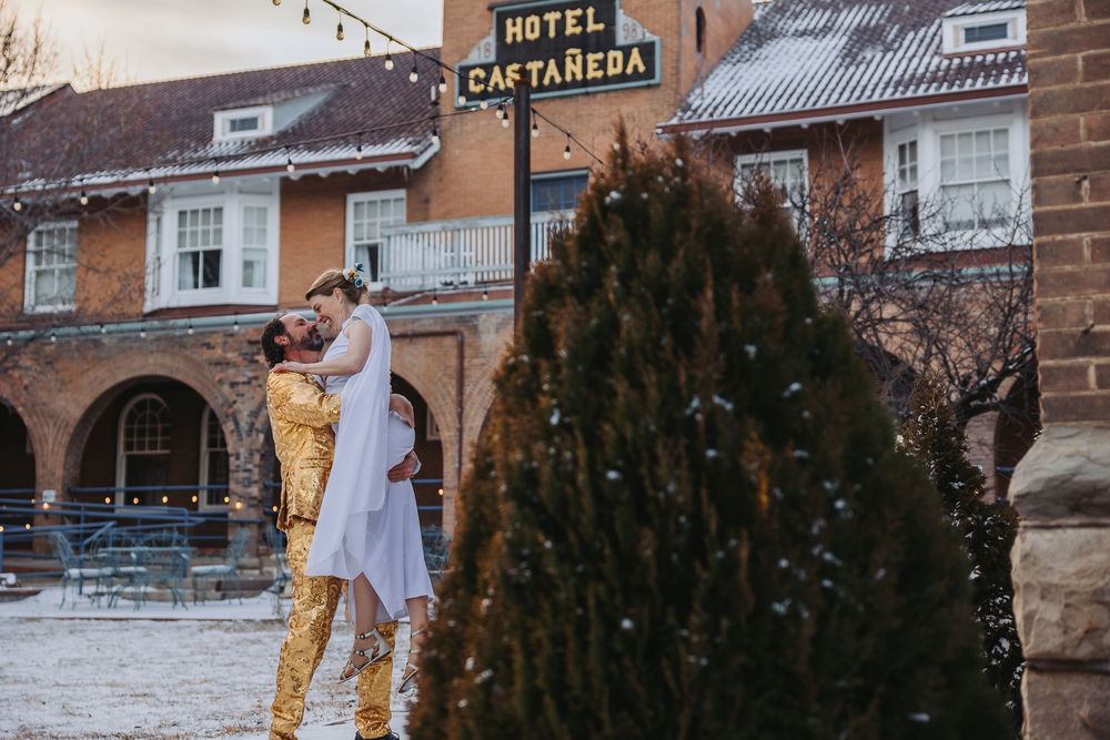 Wedding couple embraces near vintage hotel entrance with evergreen trees in winter setting.