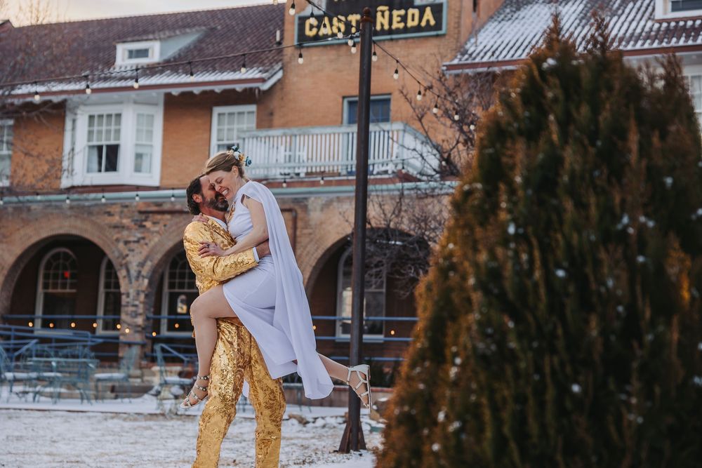 Couple in wedding attire poses in front of a rustic hotel sign and building during winter.