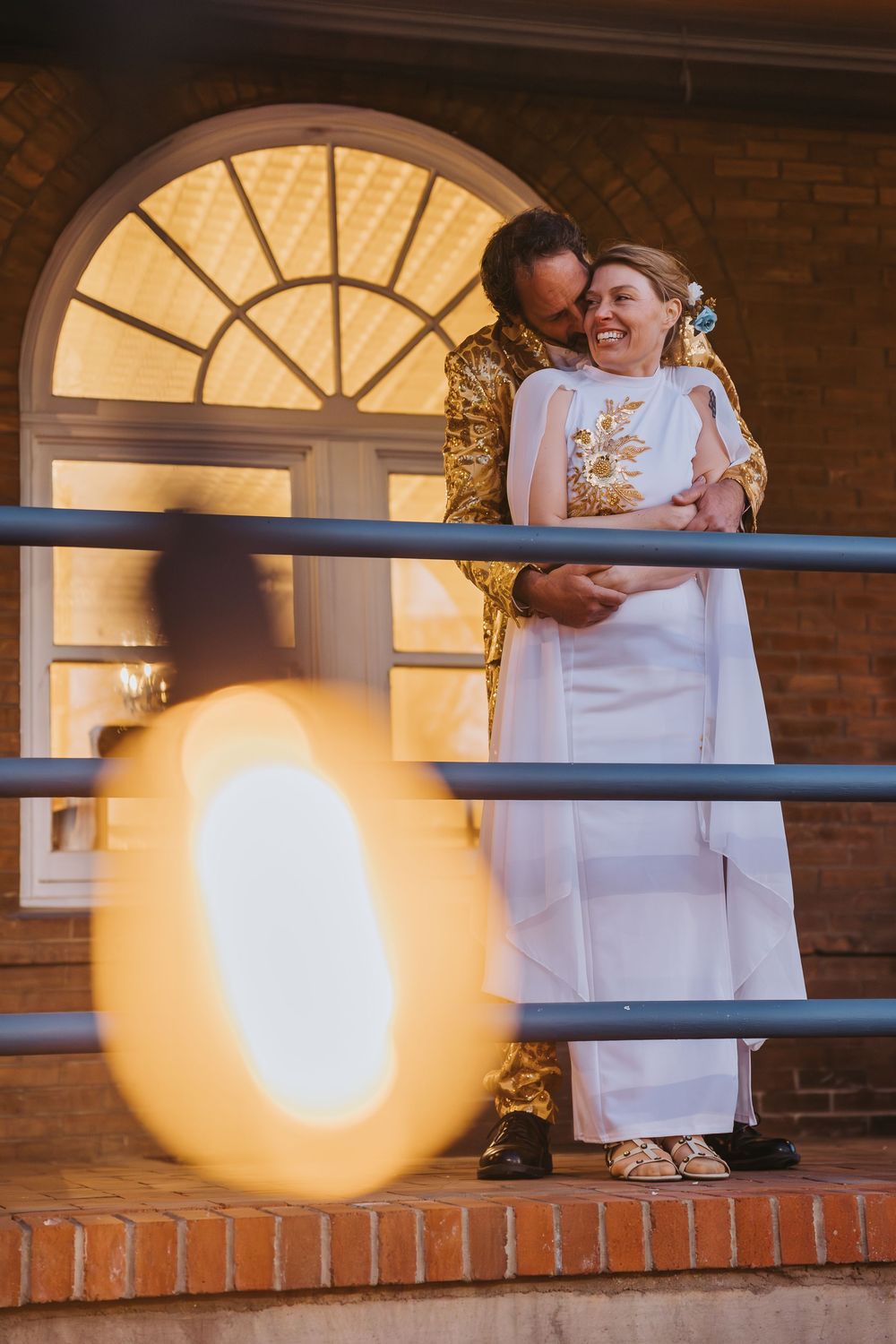 A couple shares a tender moment on a brick balcony with an illuminated arched window behind them.
