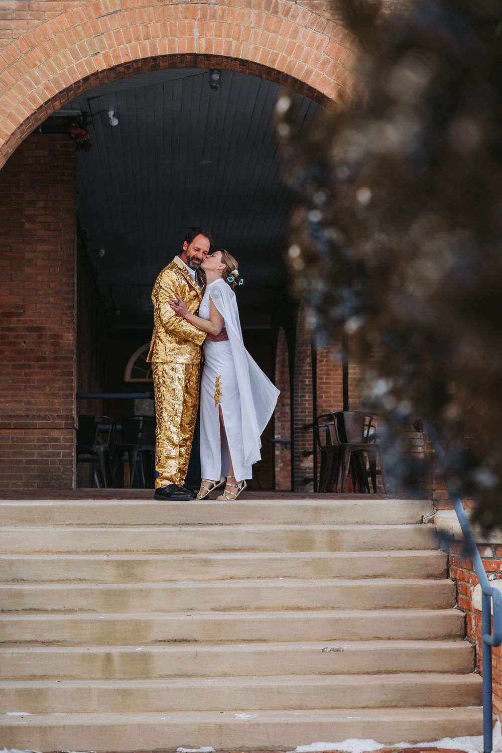 A couple in formal attire embrace on brick building steps with one person wearing a gold sequined outfit.