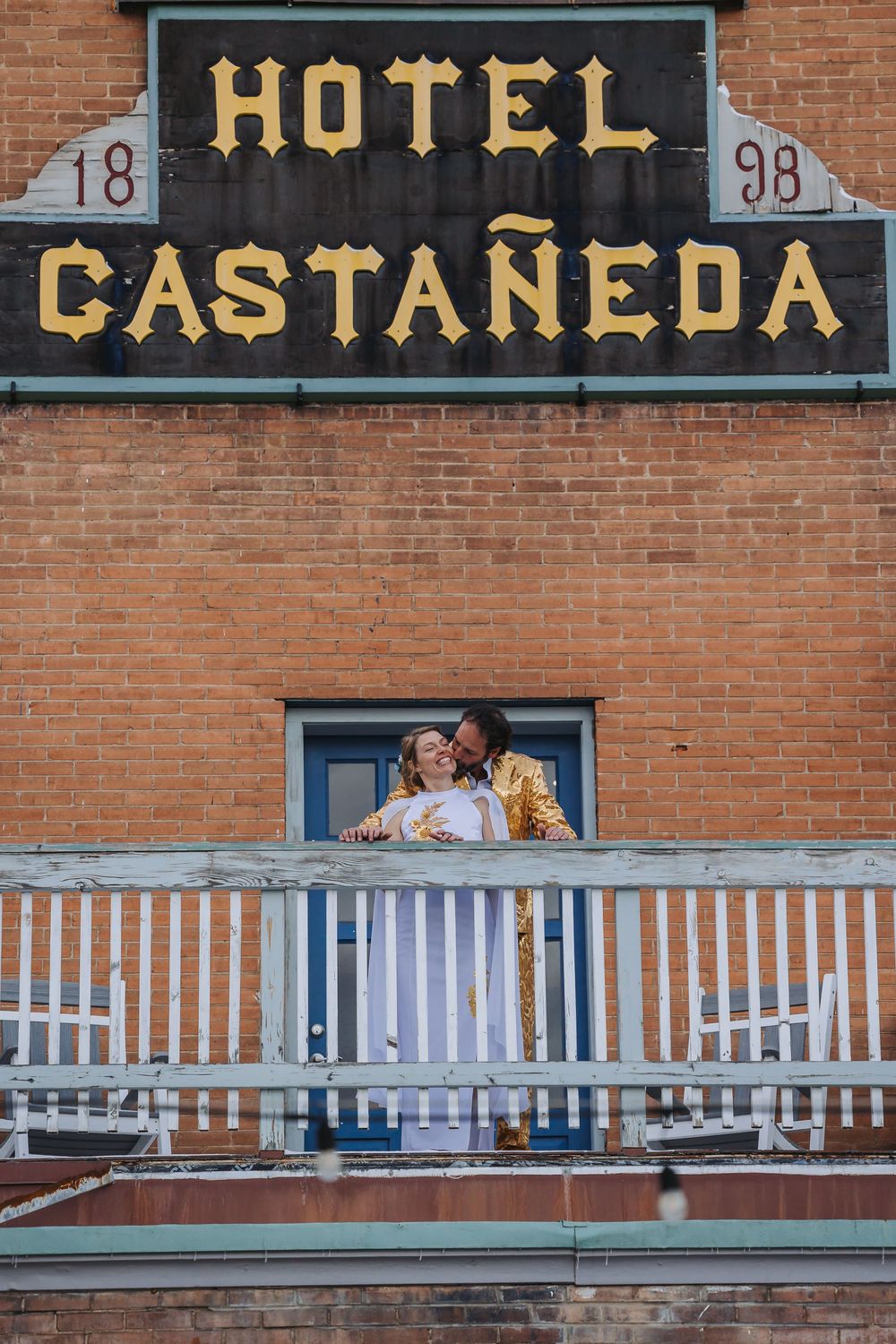 Couple embraces on a balcony at the historic Hotel Castaneda, a brick building with white railings.