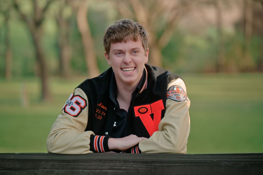 Joyful athletic senior boy smiling in letterman jacket leaning on a rustic fence in tree lined park. Vacaville, California