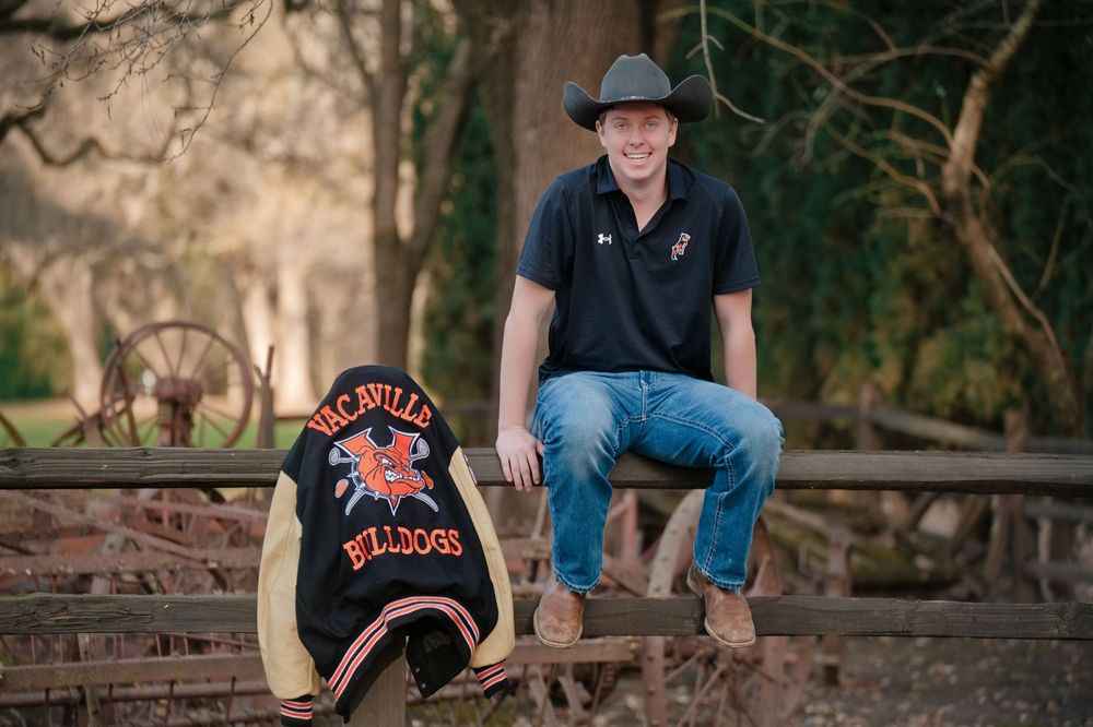 A senior young man wearing a cowboy hat sits on rustic wooden fence with his letterman jacket. Senior Portraits in Vacaville, California