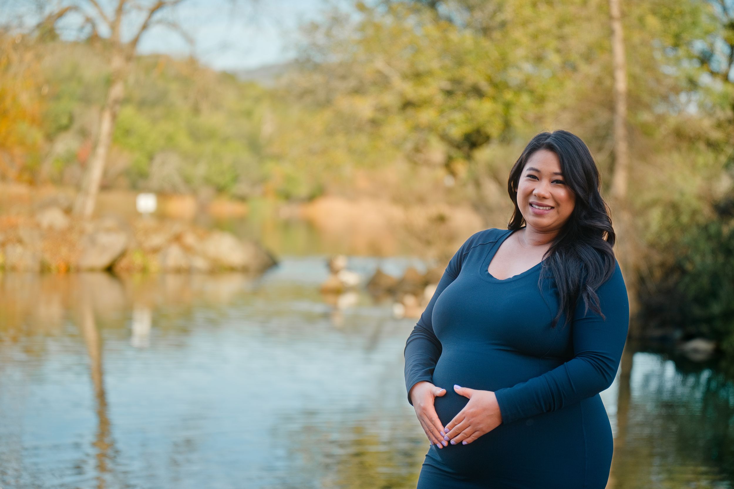 Beautiful maternity photo of mom making a heart on her baby bump in a serine park, lake side with trees. Maternity Portrait Sonoma, CA
