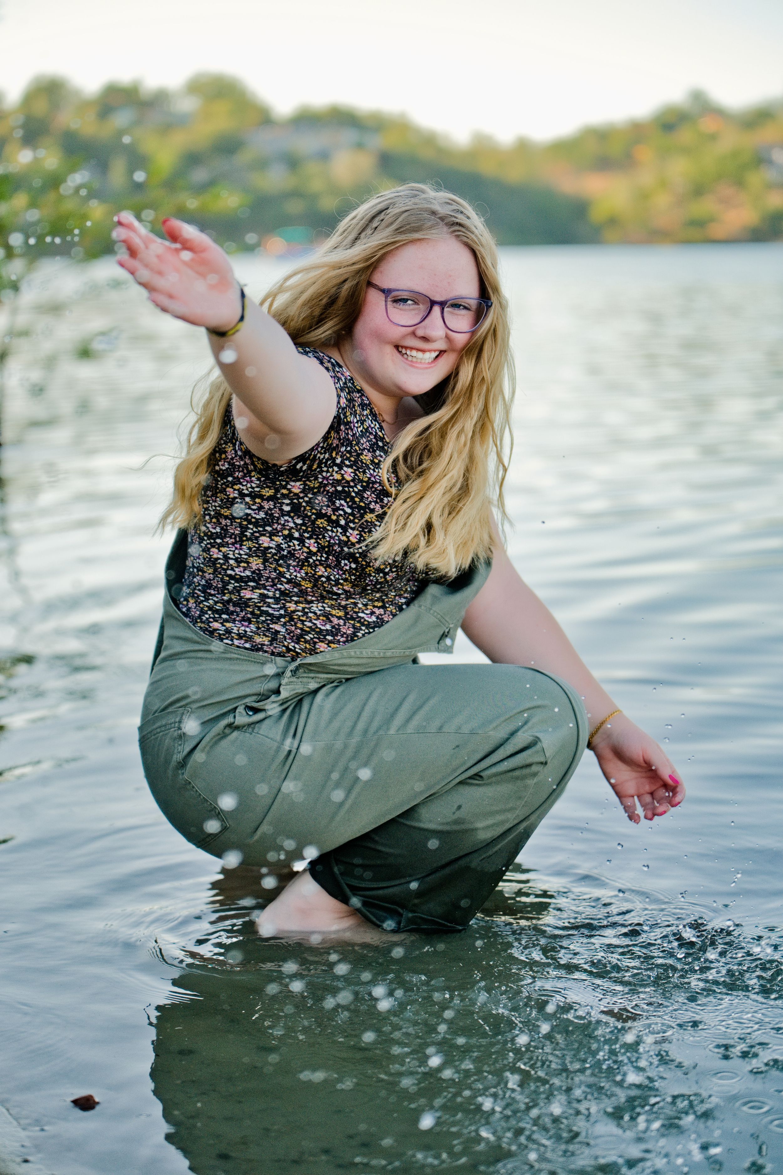 Senior girl in floral top and green pants splashes playfully in lake water during outdoor adventure photo session. Napa, California