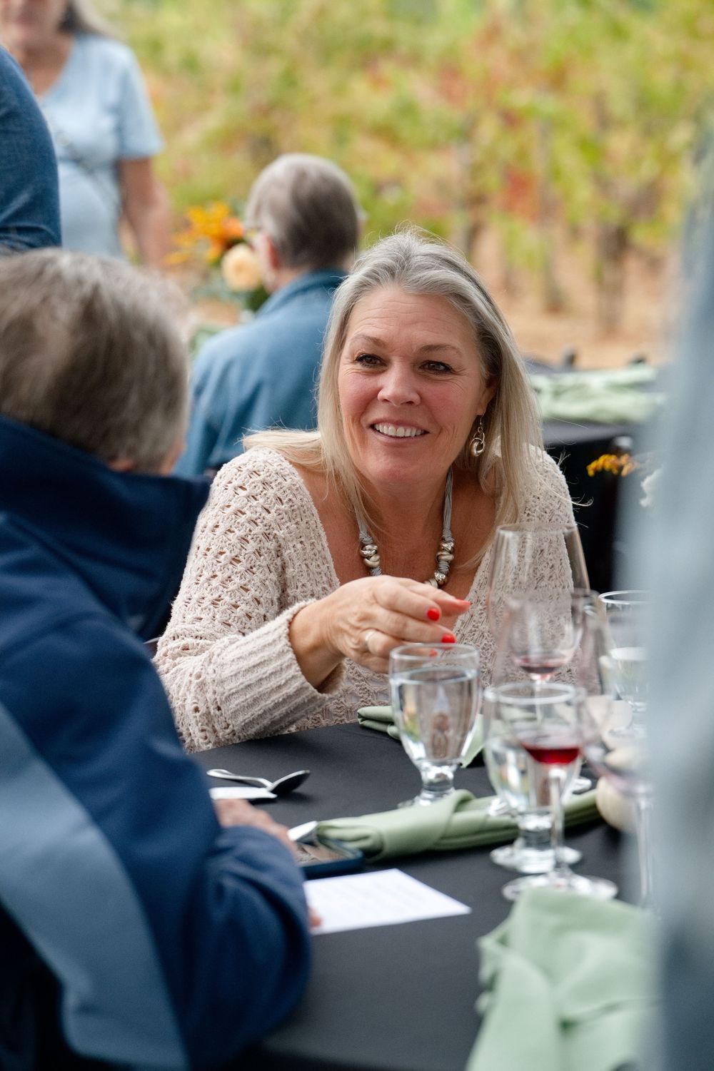 A person in a cream knit sweater sits at an outdoor dining table with friends wine glasses and place settings. Pope Valley Winery - Ca