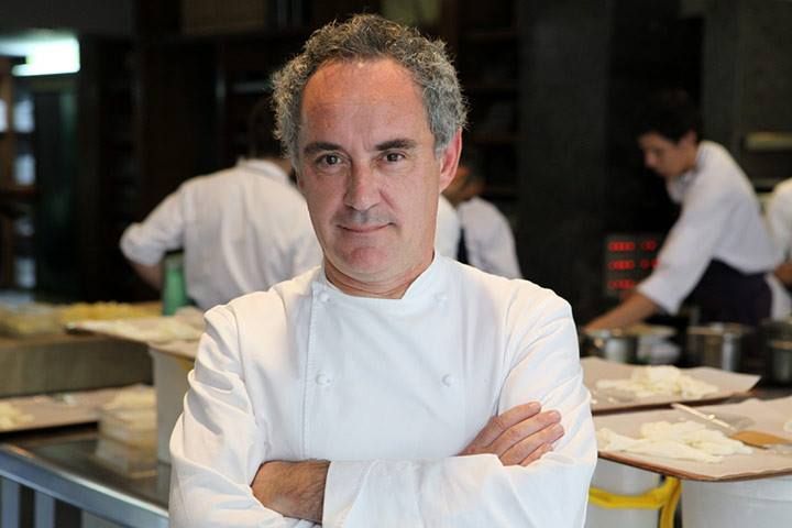 Chef in white uniform stands in professional kitchen preparing food at stainless steel counter.