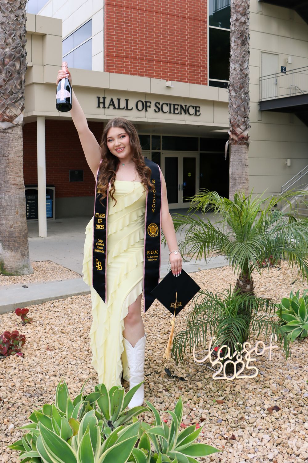 A graduate in a yellow dress and graduation stole celebrates with diploma and raised bottle outside the Hall of Science building.