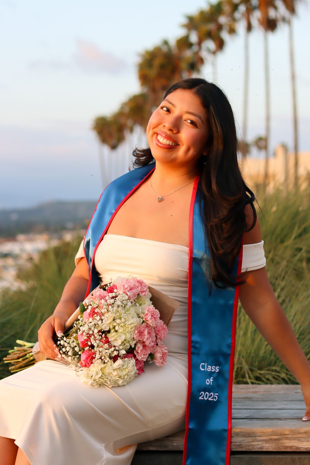 A graduate in a white dress and blue sash poses with a pink and white floral bouquet against a palm tree backdrop.