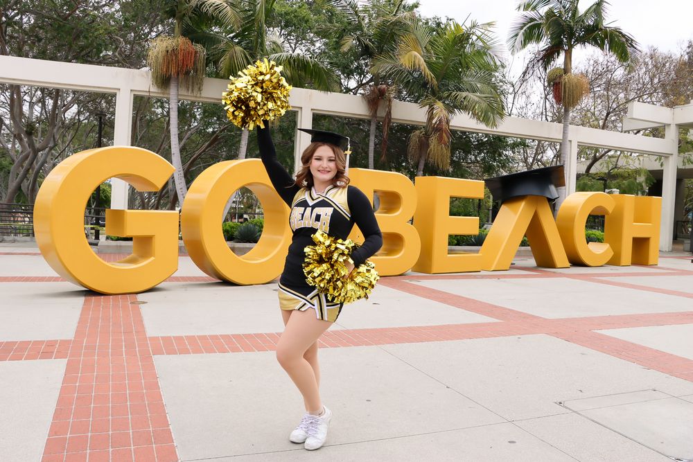 A cheerleader poses in front of large yellow 'GO BEACH' letters with palm trees in the background.