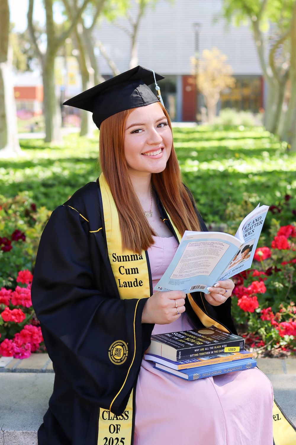 Graduate in black cap and gown poses with books and diploma against backdrop of red flowers and green foliage.