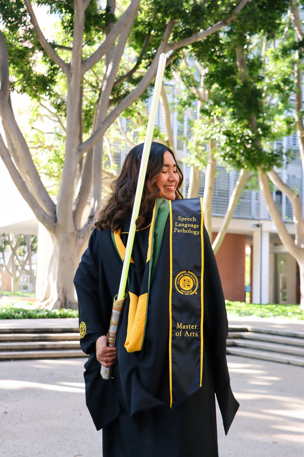 Graduate in black cap and gown with yellow stole poses for graduation photos in outdoor campus setting.