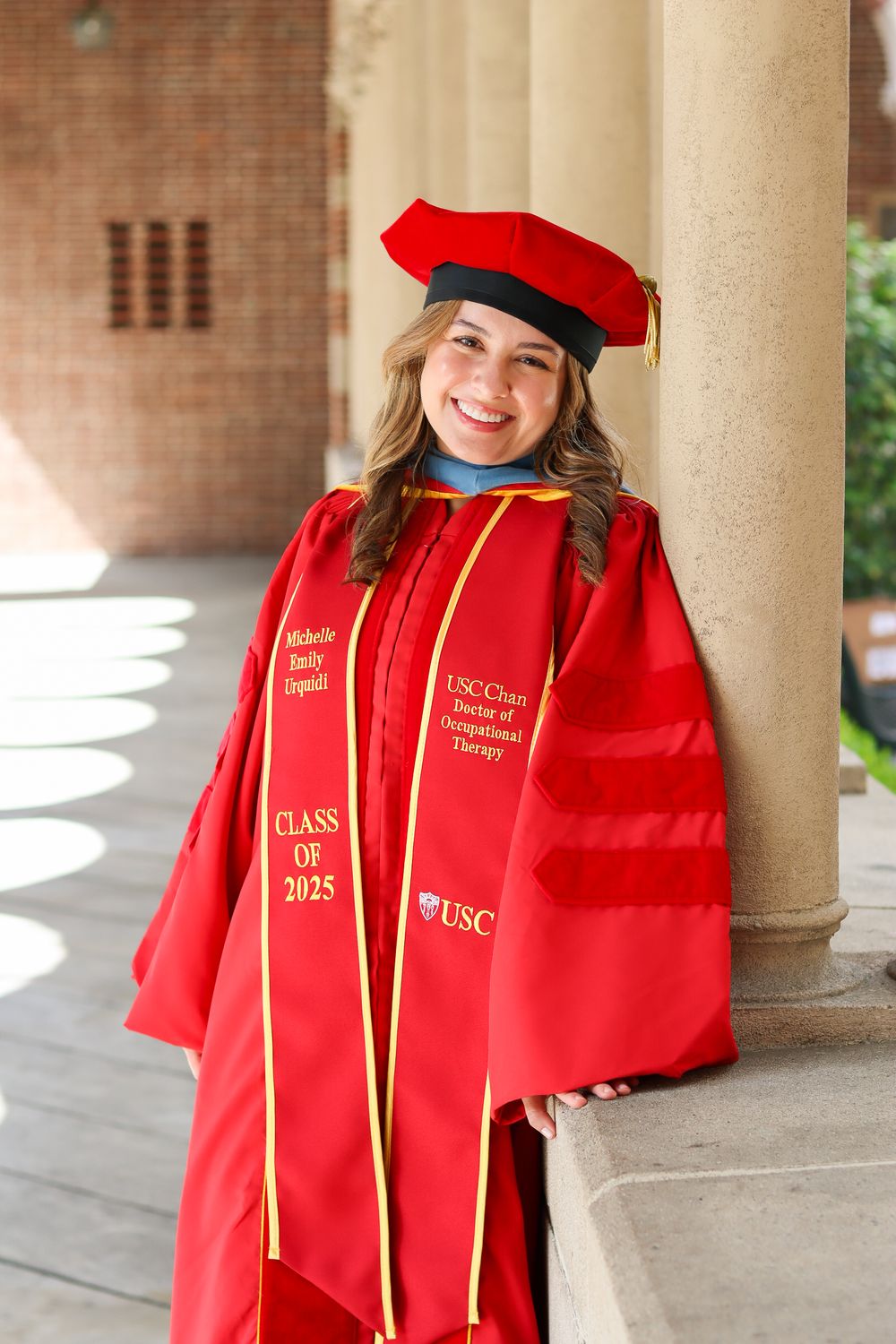 Graduate in bright red academic regalia poses for photos during commencement ceremony under brick colonnade.