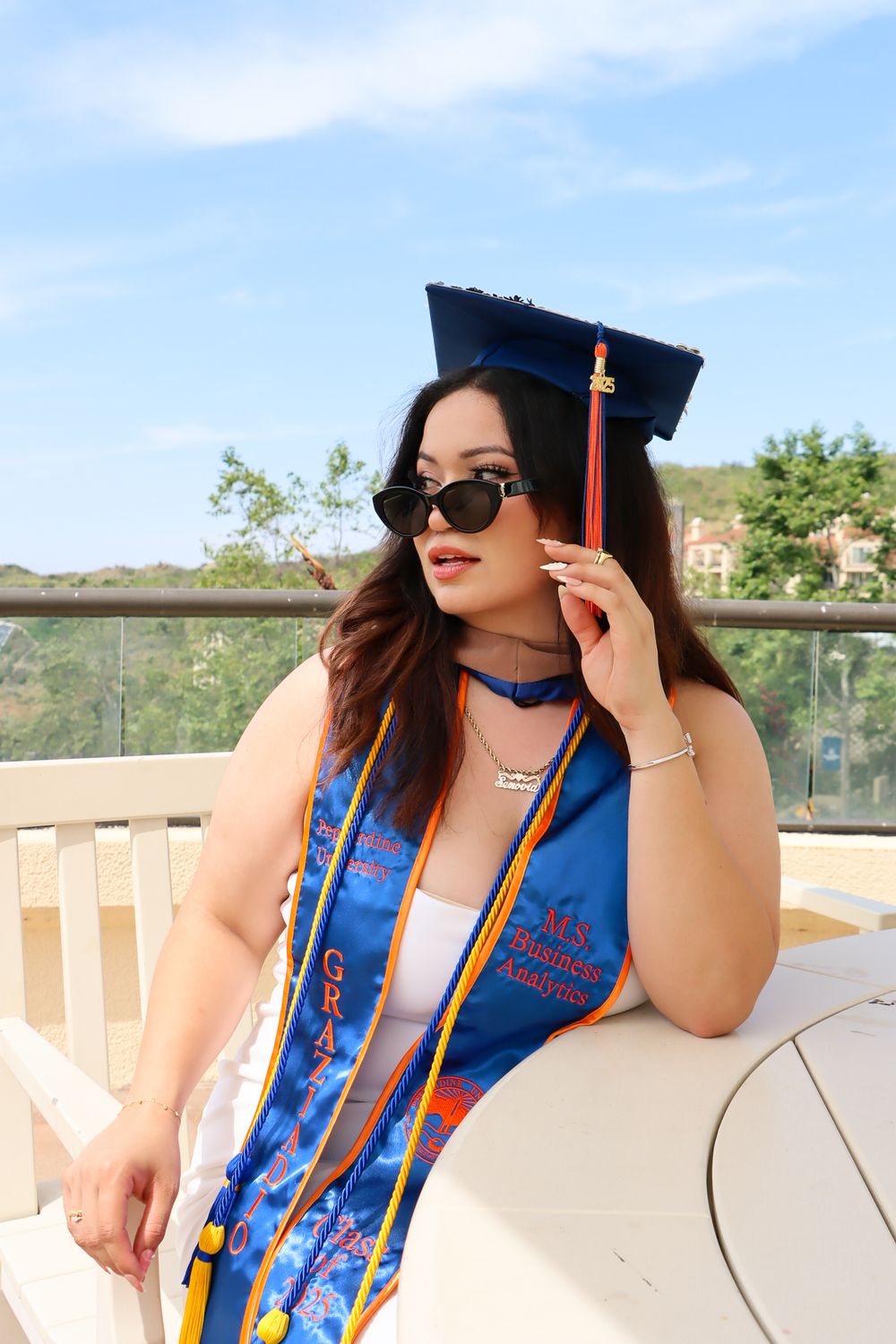 Graduate in blue cap and gown with decorative sash poses outdoors on sunny day with graduation tassel.