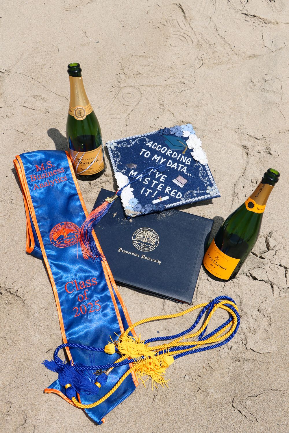 Navy blue graduation cap and gown with champagne bottle arranged on concrete surface for celebratory photo.
