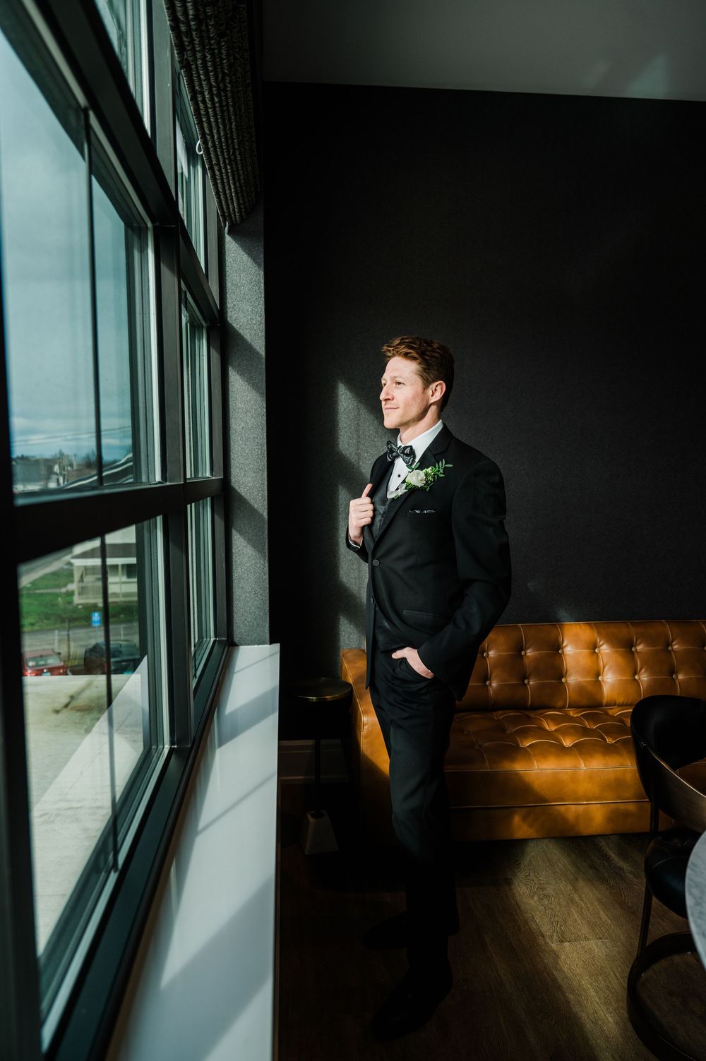 Groom in black suit with boutonniere stands contemplatively by large windows near leather couch in moody lighting.