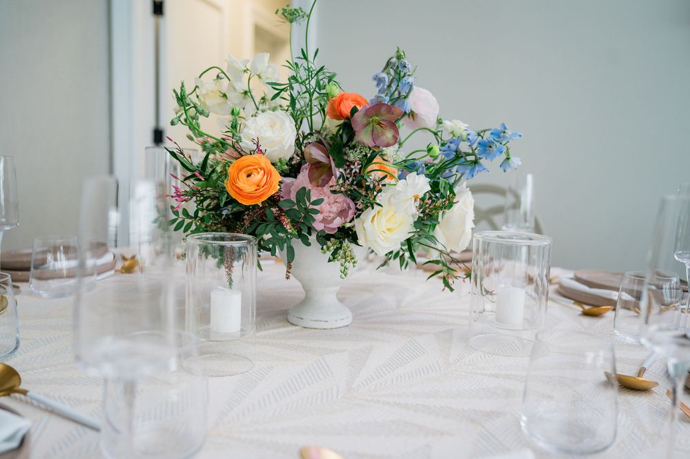Elegant floral centerpiece with orange, pink and blue flowers in a white vase on a formal dining table setting.