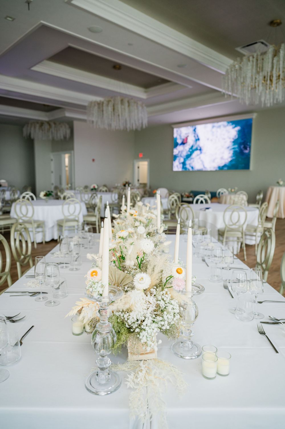 Elegant white wedding reception table with tall candles, floral arrangements and crystal chandeliers overhead.