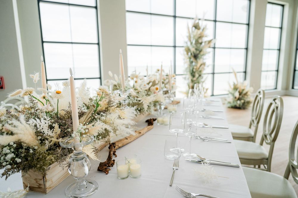 Elegant white and gold wedding reception tables with floral centerpieces in a bright modern venue with large windows.