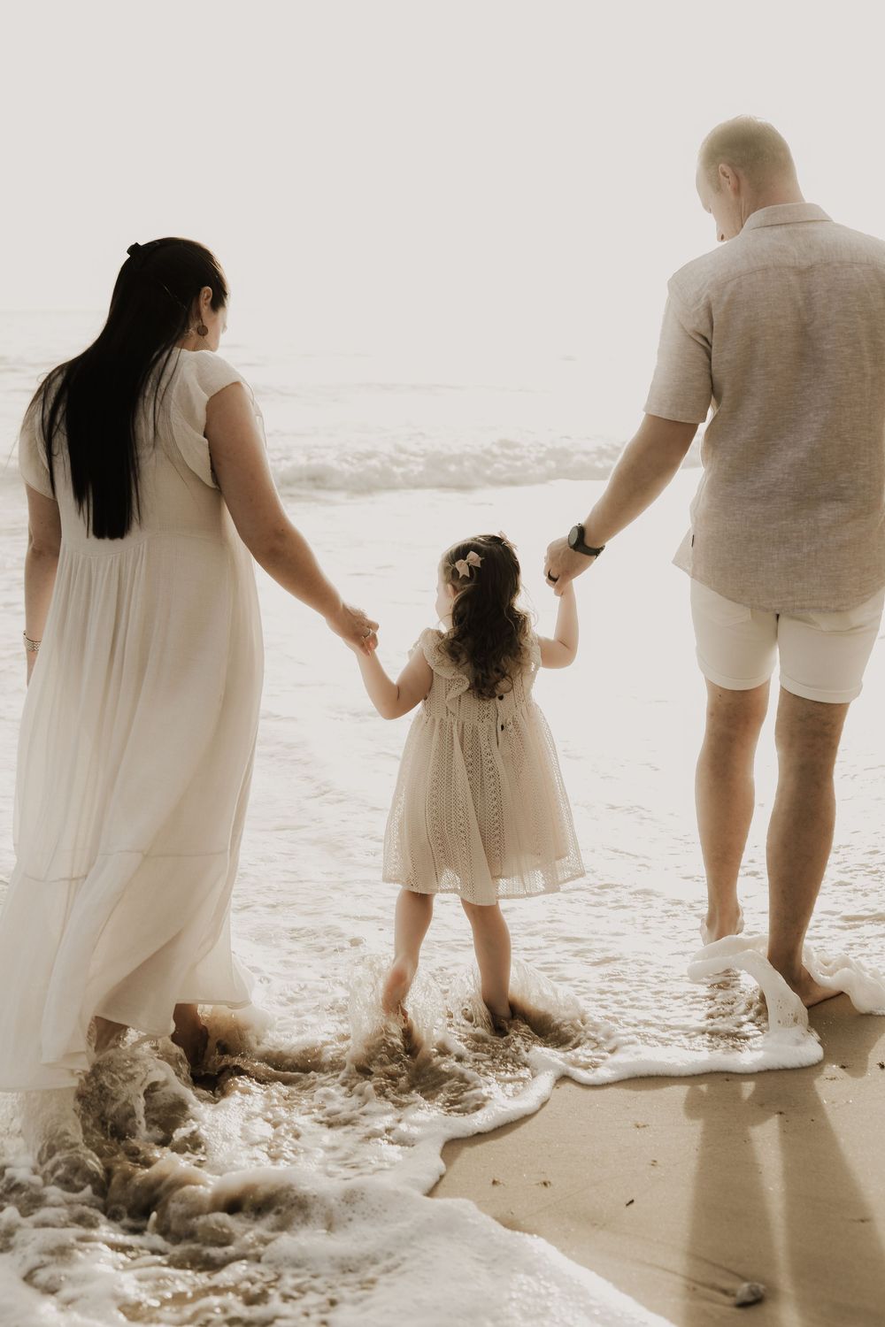 Family of three holding hands at the beach, parents in white outfits with young daughter in a white dress by ocean waves.