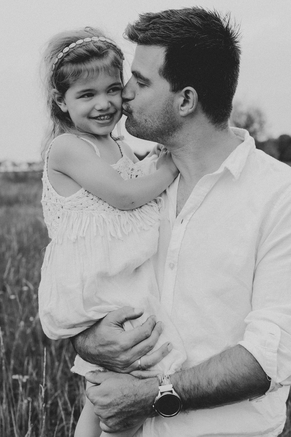 Black and white photo of a father kissing his smiling young daughter on the cheek outdoors in a field.