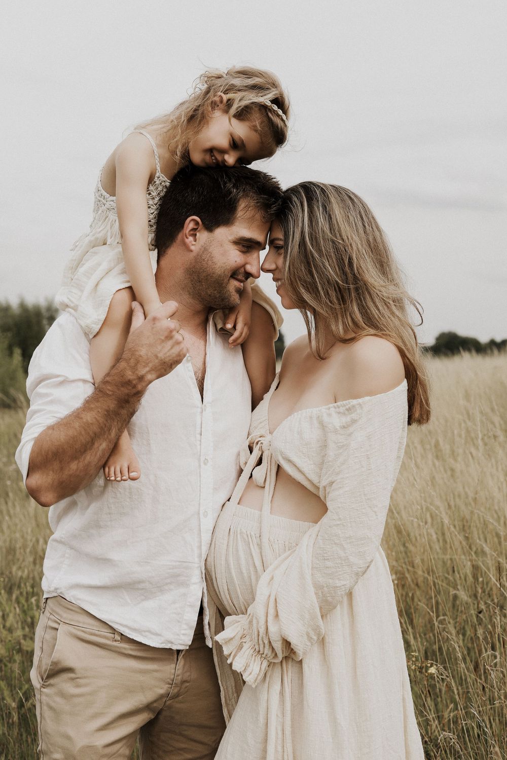 A happy family of three poses in a golden field, with a young girl sitting on her father's shoulders.