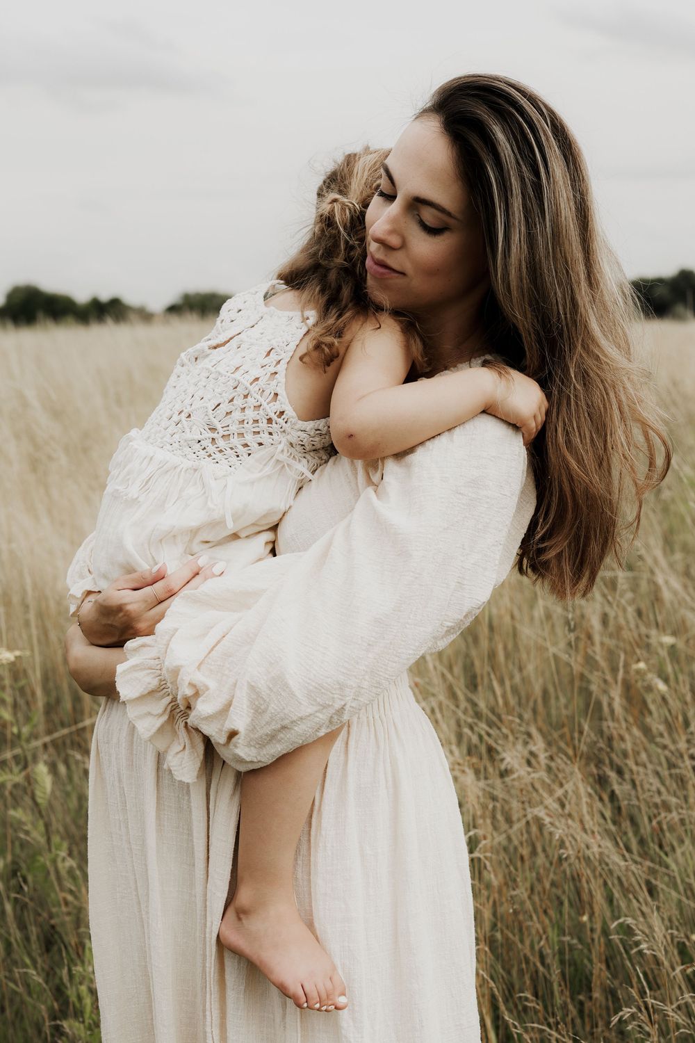 A mother in a white dress tenderly holds her young child in a golden grass field.