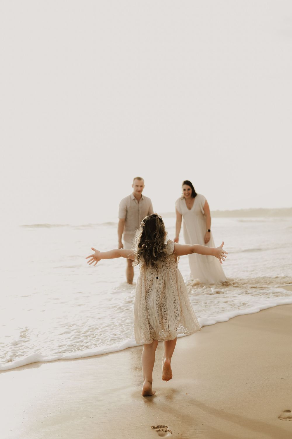 A child in a white dress runs toward a couple on a sunlit beach, arms outstretched in a joyful family moment.