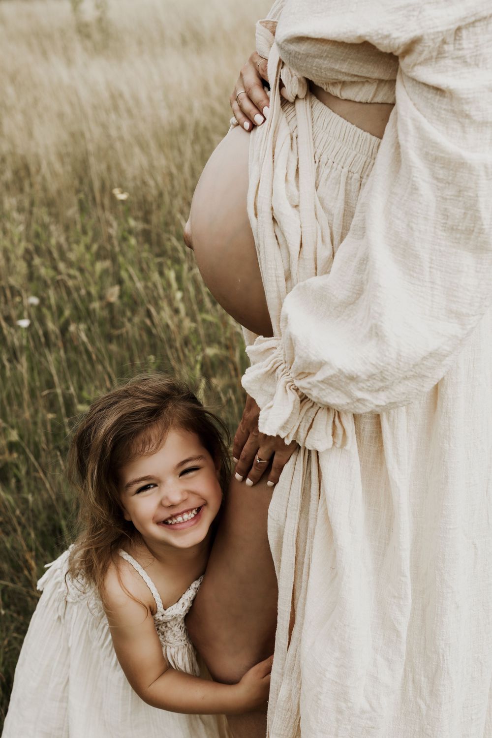 A smiling little girl in a white dress hugs a pregnant woman's belly in a sunlit field.