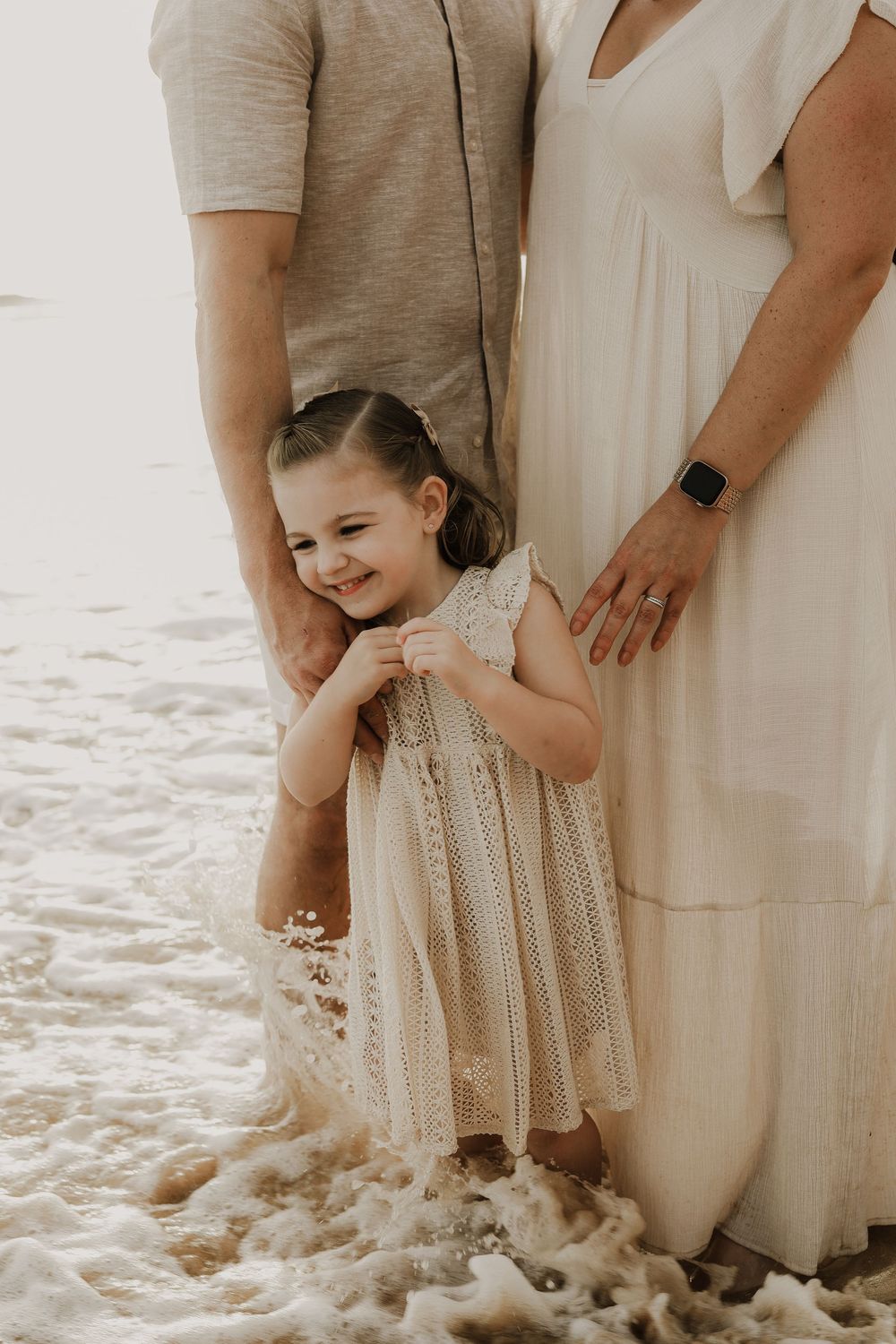 A smiling toddler girl in a lace dress stands between her parents on a beach as ocean waves wash over her feet.