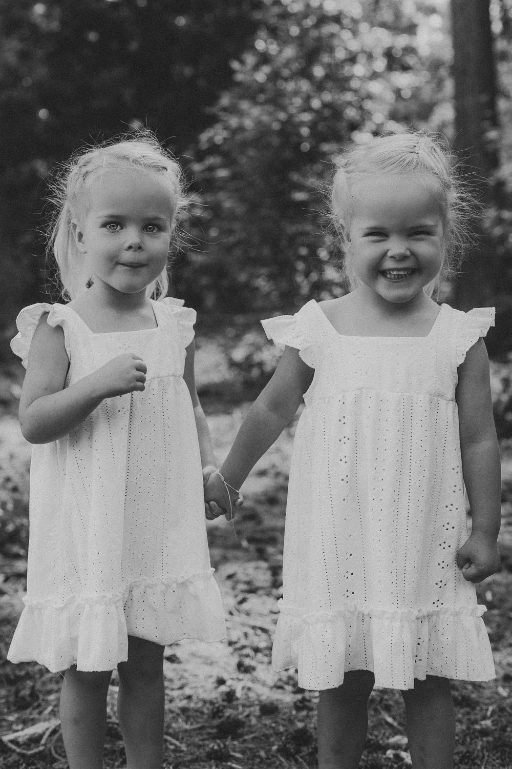 Two young blonde sisters in matching white dresses smiling in a black and white outdoor portrait.