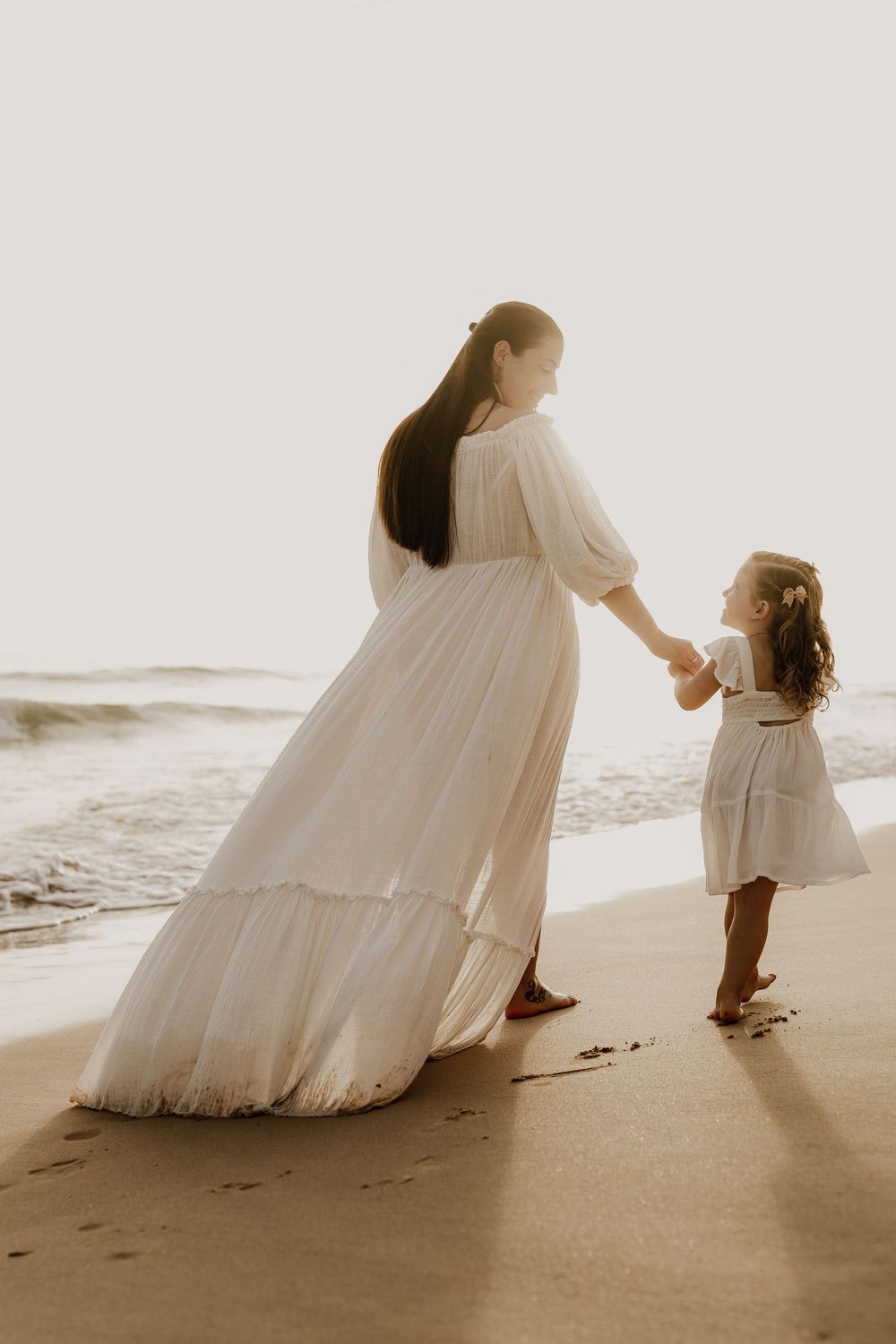 A mother and daughter in white dresses hold hands on a sunlit beach, sharing a tender moment by the ocean waves.