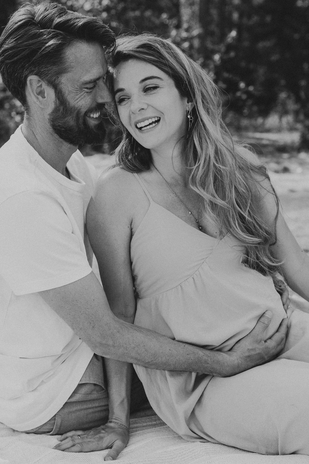 Black and white maternity photo of a laughing pregnant woman and her partner sitting together outdoors.