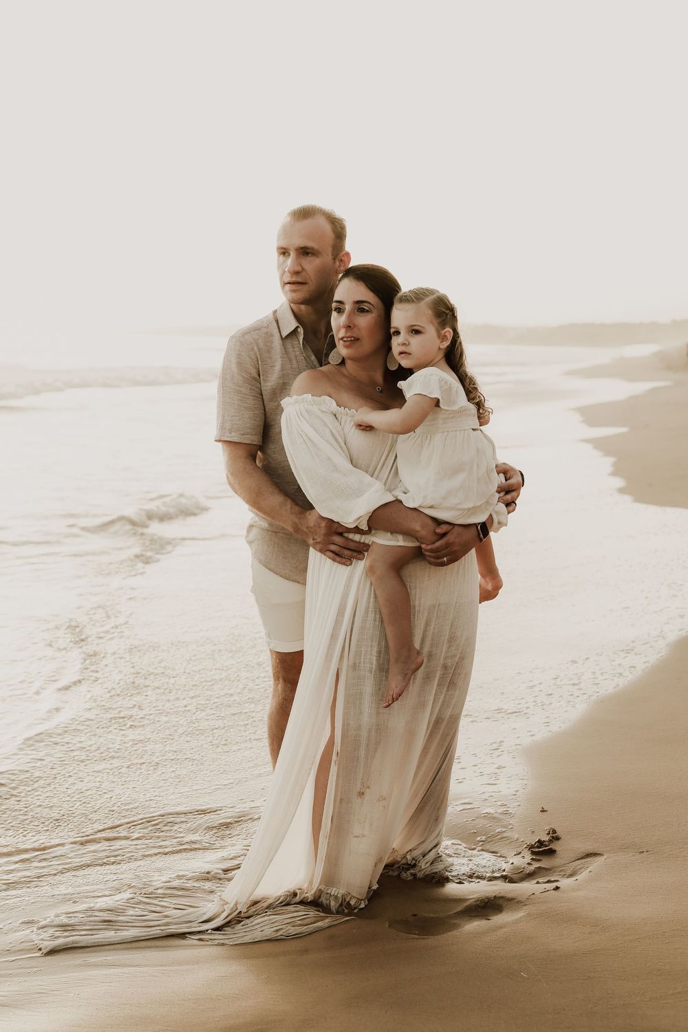 Family of three posing on a sandy beach at sunset, mother in white dress holding toddler, father beside them.